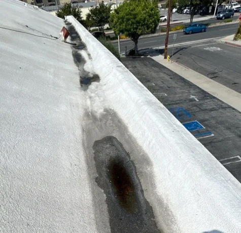 View from a rooftop showing a long dark wet stain along the white roof edge, with a street and parked cars visible below.